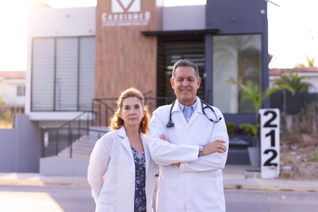 Médico cardiólogo revisando a un paciente adulto en un consultorio, con estetoscopio y equipo de monitoreo cardíaco al fondo