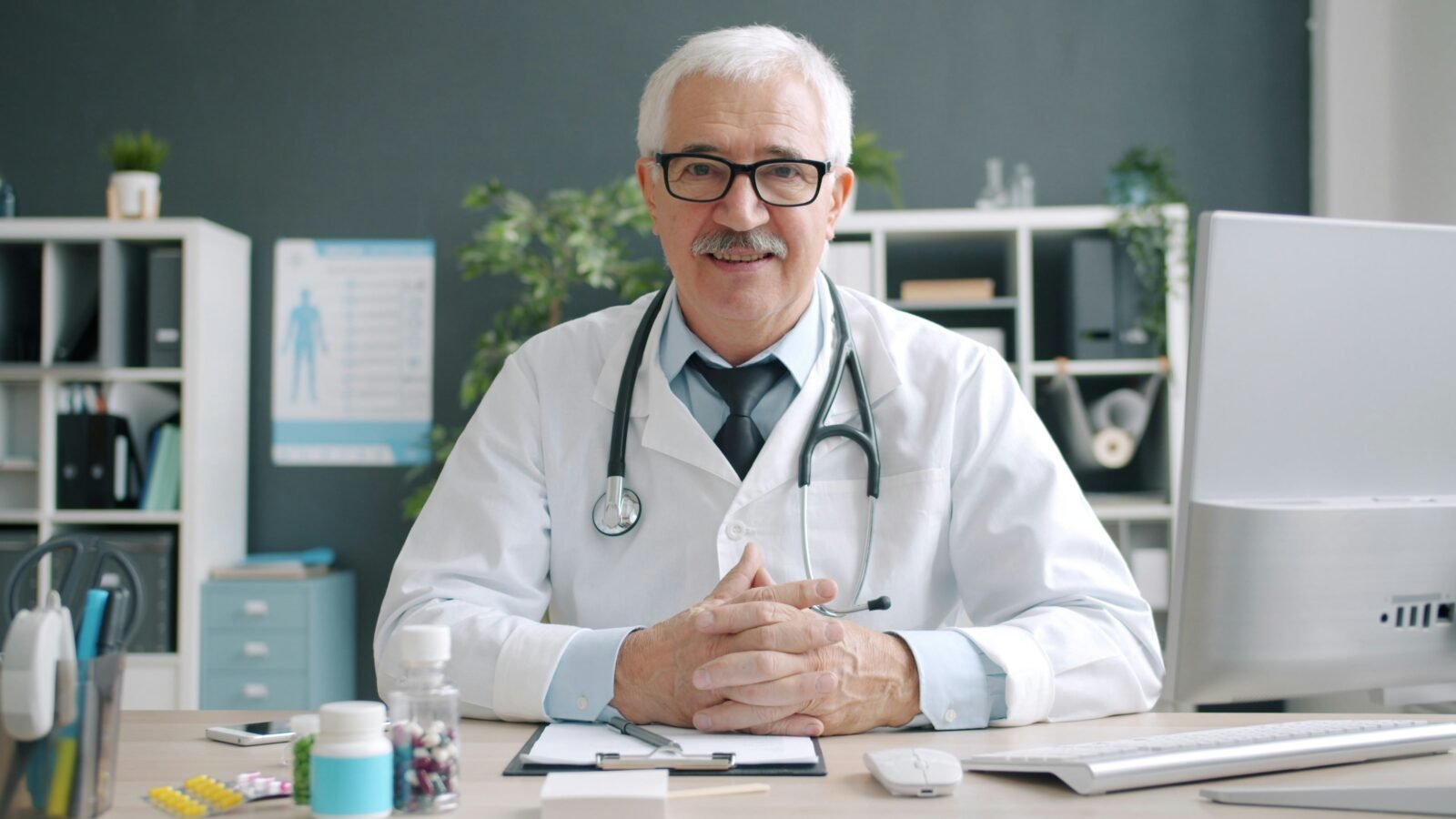 Médico canoso mirando a cámara, sentado en su consultorio con las manos apoyadas sobre el escritorio