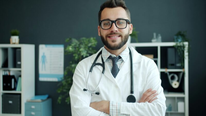 Médico de guardapolvo blanco mirando a cámara en su consultorio